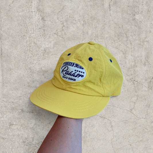 Yellow vintage style baseball cap with navy blue oval Scandinavian logo patch on the front, held by hand against a grey textured background