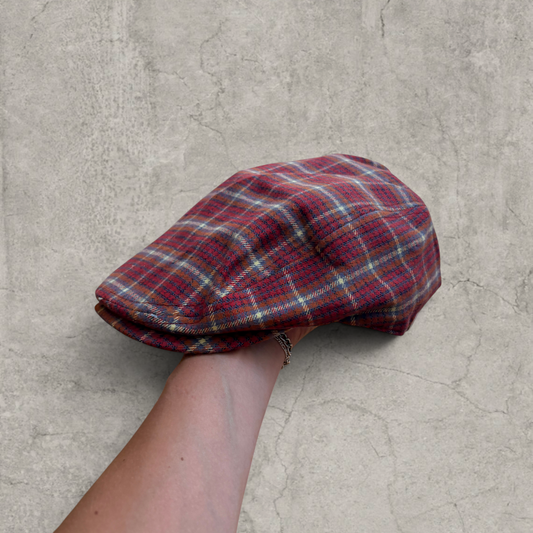 Plaid flat peak beret cap in burgundy red held by hand against a grey concrete background, showing front and top view