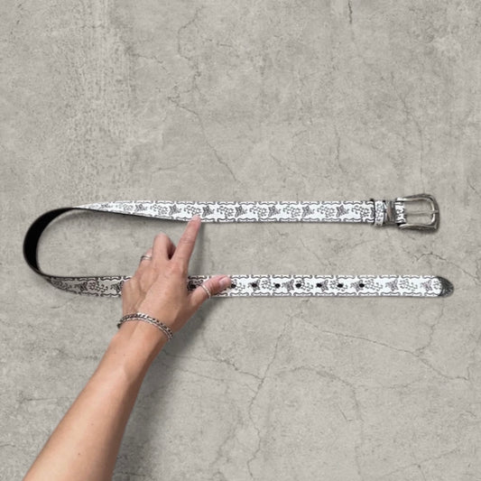 White vintage style faux leather western cowboy belt with silver trim laid flat on grey surface, hand pointing at pattern