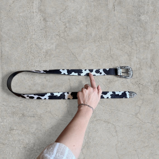 White and black cow print faux leather western cowboy belt laid flat with silver engraved buckle and tip, hand pointing to centre