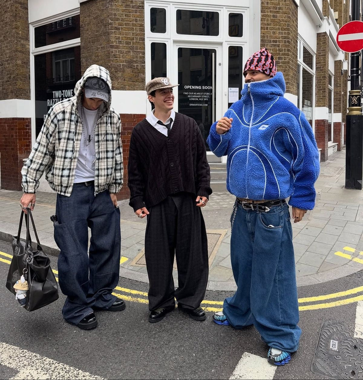 Three men standing on a city street in front of a brick building corner. The person on the left wears a white and black plaid hoodie over a white shirt with dark blue baggy jeans and holds a black bag.
