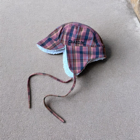 Checkered navy blue pink brown trapper ear flap hat with fluffy white lining on beige surface