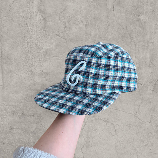 Blue, turquoise and black checkered plaid baseball cap with white logo held in hand against a grey textured wall