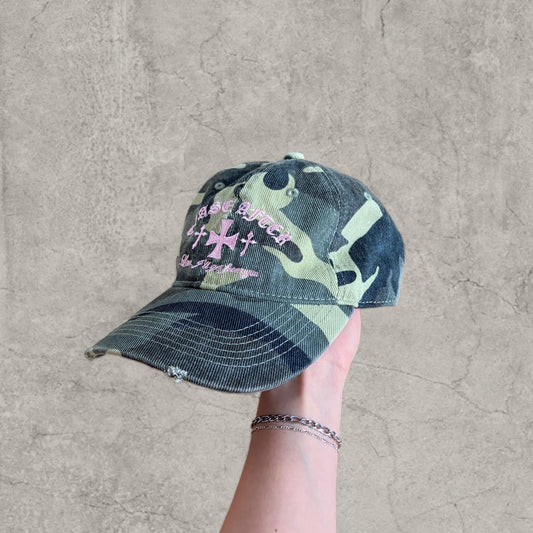 Hand holding vintage style baseball cap with khaki green camouflage print and pink gothic logo