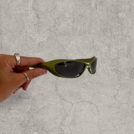Khaki green wrap-around sunglasses with black lenses held in hand against a grey wall