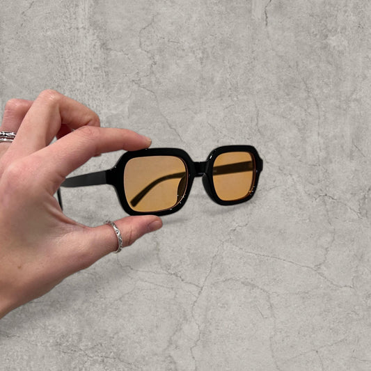 Black framed glasses with orange tinted square lenses held in hand against grey background