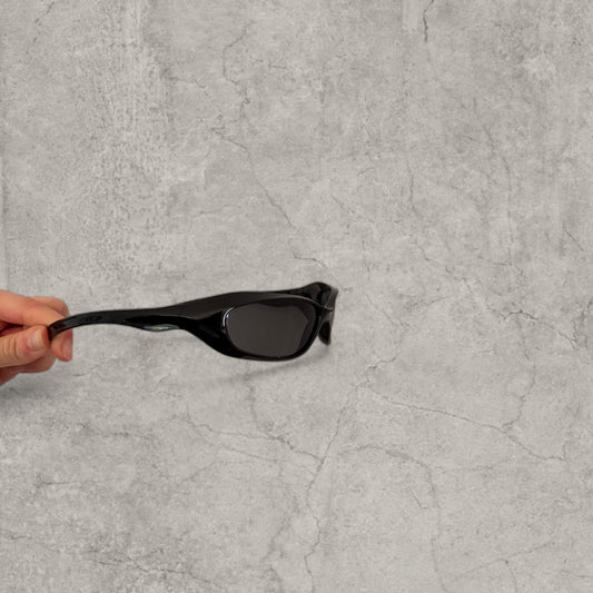 Black wrap-around plastic sunglasses with black lenses held at one arm side on grey stone background