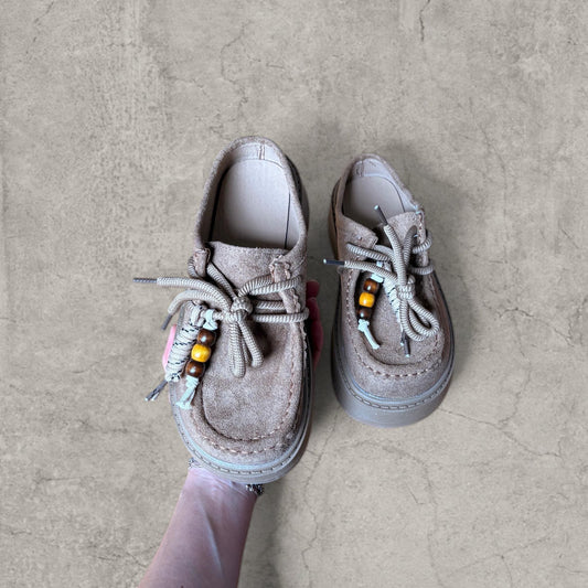 Brown lace-up mule clog shoe with tassels and bead detailing held above a concrete floor