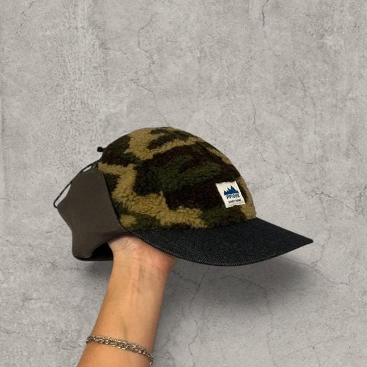 Trapper Russian cap with camo fleece crown and navy curved brim held against a grey textured background, showing side and front strap