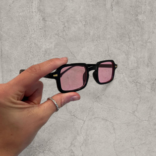 Hand holding black frame glasses with square pink tinted lenses over a grey textured background