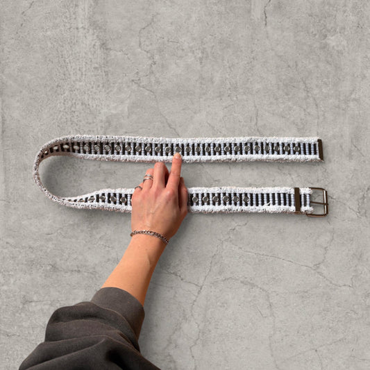 Distressed denim belt in black and white with grey eyelets laid flat on concrete floor showing hand pointing to eyelet