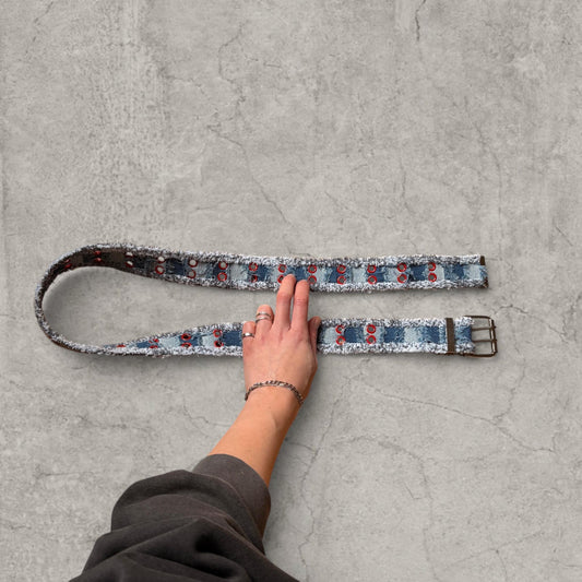 Distressed denim belt in navy and light blue stripes with red eyelets laid flat on concrete surface