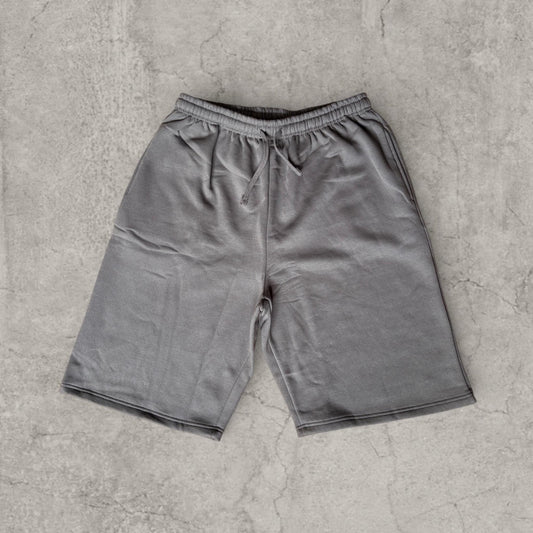 Grey baggy wide-leg long jersey shorts laid flat on concrete surface, showing front drawcord waist and side pockets