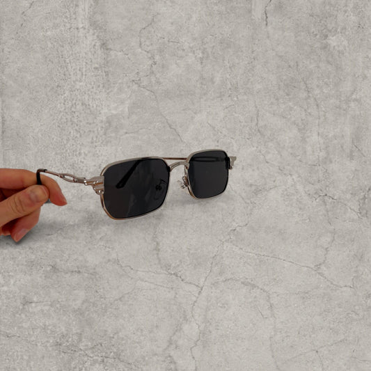 Silver grey Insane sunglasses with black square lenses shown handheld against a grey surface