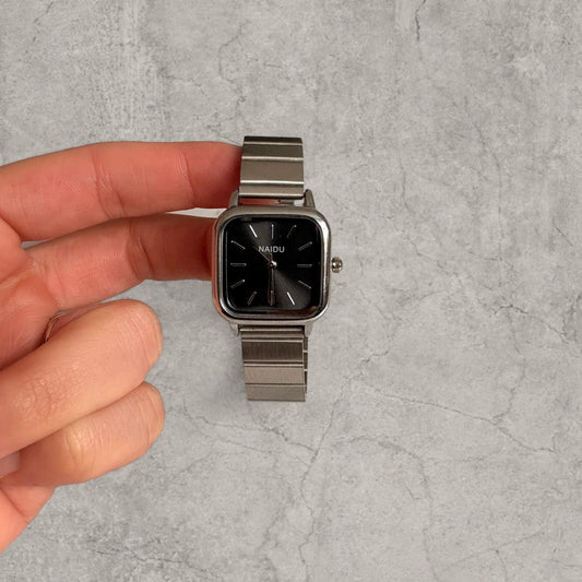Hand holding metal watch with square grey face showing front dial with silver markers