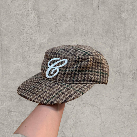 Vintage style baseball cap with brown beige houndstooth plaid and white logo held against grey cracked wall