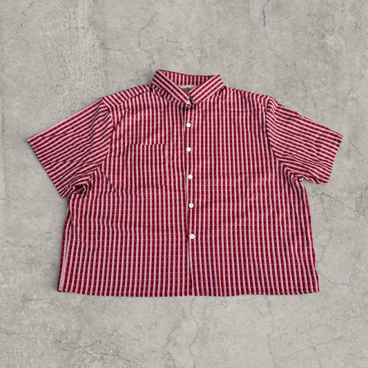 Boxy short-sleeve shirt in red and white vertical stripes laid flat on a concrete floor, front view showing collar and buttons