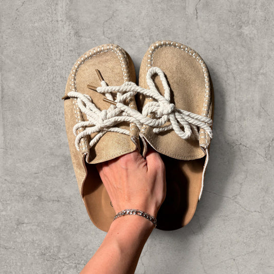 Pair of beige brown suede lace-up mule shoes with cream rope laces held in hand against concrete background