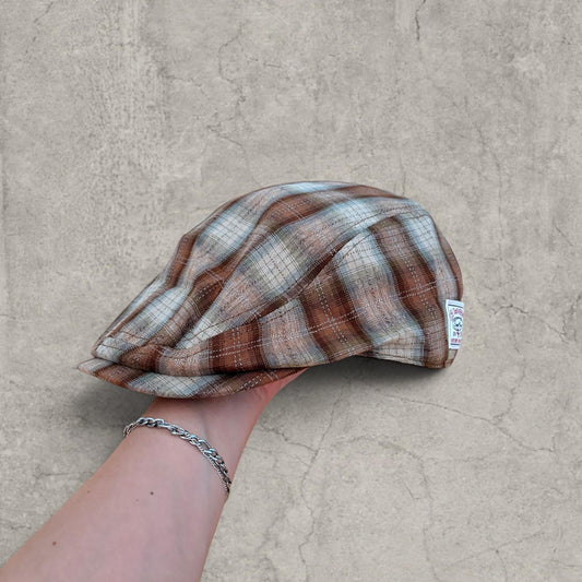 Flat peak beret cap in brown beige checkered plaid held by hand against a stone background